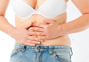 A person wearing a white bra and blue jeans, with both hands resting on their stomach. The image focuses on the torso area, highlighting a natural pose and expression related to discomfort. from dosing wiith semaglutides and tirzepatide early in treatment