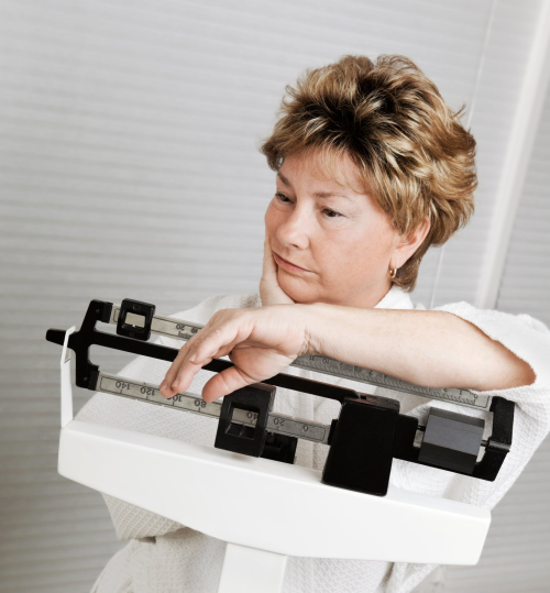 A woman in a white robe thoughtfully adjusting a balance scale in a bright, minimalistic room. The scale displays weights, reflecting her focus on weight management as she considers the effects of microdosing GLP-1 medications for weight loss and maintenance.