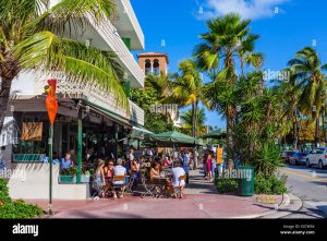 A lively outdoor café scene with people dining at tables shaded by large umbrellas. The surrounding area features palm trees and a clear blue sky, with additional pedestrians walking along the sidewalk.