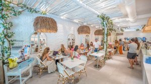 A bright, trendy café interior featuring white brick walls and large straw pendant lights. Groups of people are seated at small tables, engaged in conversation. Green plants adorn the space, creating a lively atmosphere.