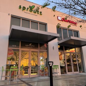 The exterior of Sproutz Healthy Kitchen, featuring large glass windows and a welcoming entrance. Next to it is the sign for a restaurant named 'China Natural.' The scene is vibrant with outdoor seating and soft lighting.