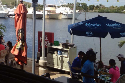 A lively outdoor dining scene by a waterfront, featuring diners at tables near a grill station. An umbrella with 'Modelo' branding shades the guests, with a view of boats docked in the water. This scene captures a casual dining atmosphere in Cutler Bay/Homestead, Fl.