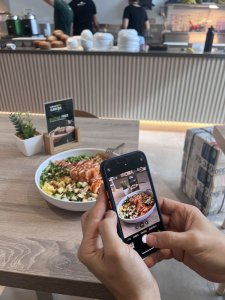 A person holding a smartphone, capturing a photo of a colorful salad bowl filled with vegetables and grilled chicken, on a wooden table in a casual dining space. In the background, staff members are visible in a modern eatery.