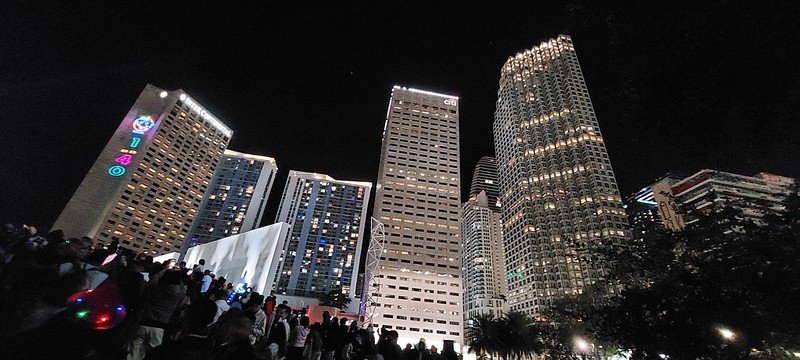A vibrant nighttime cityscape featuring tall skyscrapers illuminated by lights. A crowd of people stands in the foreground, with a few individuals wearing colorful accessories, enjoying the lively atmosphere. OF bRICKELEL aVE AREA OF MIAMI