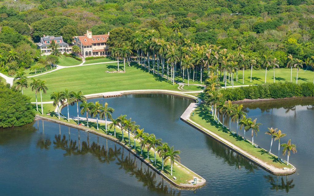 Aerial view of a lush, green estate with a large house surrounded by palm trees. Curved pathways and manicured lawns extend towards a serene, dark waterway lined with more palm trees, creating a tranquil landscape.