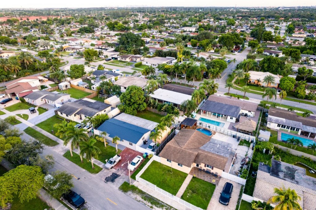 An aerial view of a suburban neighborhood in hialeah, FL, showcasing a mix of single-family homes with well-maintained lawns and palm trees lining the streets. Some homes feature swimming pools in their backyards, illustrating a vibrant community.