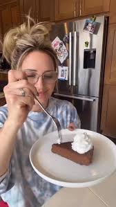 A woman wearing glasses holds a fork poised above a slice of chocolate dessert topped with whipped cream on a white plate. In the background, a kitchen with wooden cabinets and a refrigerator can be seen. This moment illustrates enjoyment of food and food noises.