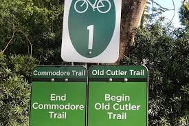 A green trail sign indicating the transition from the Commodore Trail to the Old Cutler Trail, with instructions to end the Commodore Trail and begin the Old Cutler Trail. A bicycle symbol is featured above the trail number 1.
