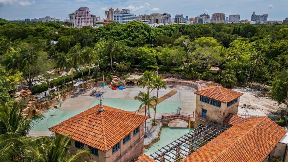 Aerial view of a large outdoor space featuring a colorful playground surrounded by lush green trees, with a city skyline in the background. The area appears sunny and inviting, showcasing both nature and urban architecture. This setting may be appealing for families, highlighting how GLP-1 Medications Help Coral Gables Residents.